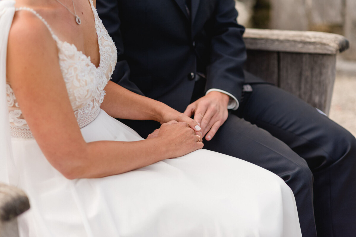 Ein Paar im Hochzeitskleid sitzt auf einer Holzbank und hält sich an den Händen.