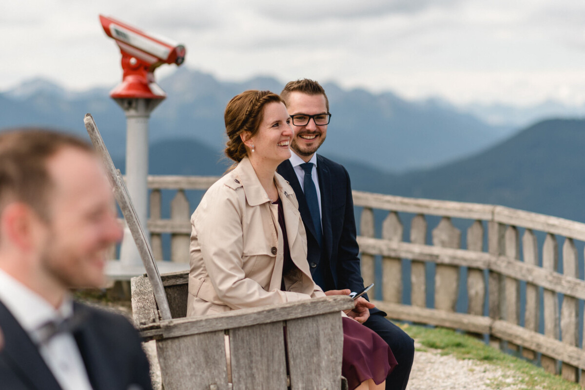 Zwei Personen sitzen lächelnd auf einer Holzbank mit Bergen im Hintergrund. In der Nähe steht ein rotes Fernglas.