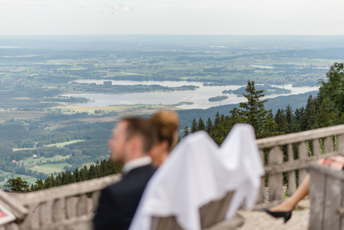 Ein Paar sitzt auf einer Bank und blickt auf eine weite Landschaft mit Feldern, Wäldern und einem entfernten Fluss oder See. Ihre Gesichter sind verschwommen und der Blick ist auf die malerische Aussicht gerichtet.
