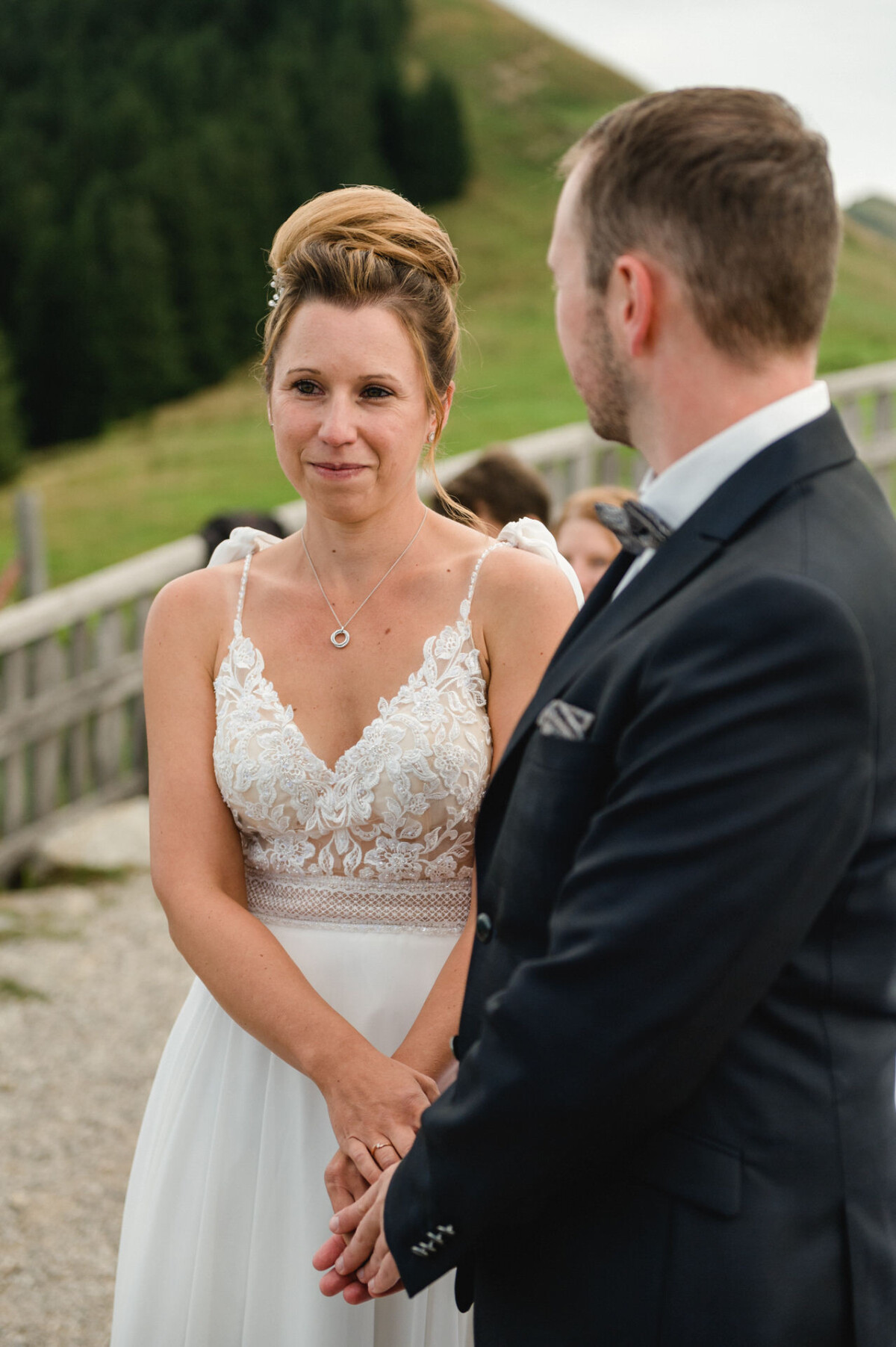 Braut und Bräutigam stehen bei einer Zeremonie im Freien zusammen. Die Braut trägt ein weißes Spitzenkleid und der Bräutigam einen dunklen Anzug. Im Hintergrund sind ein Holzzaun und Grünpflanzen zu sehen.