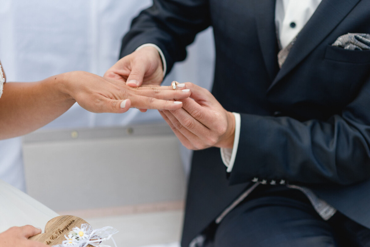 Eine Person im Anzug steckt während einer Zeremonie einer anderen Person einen Ring an den Finger.