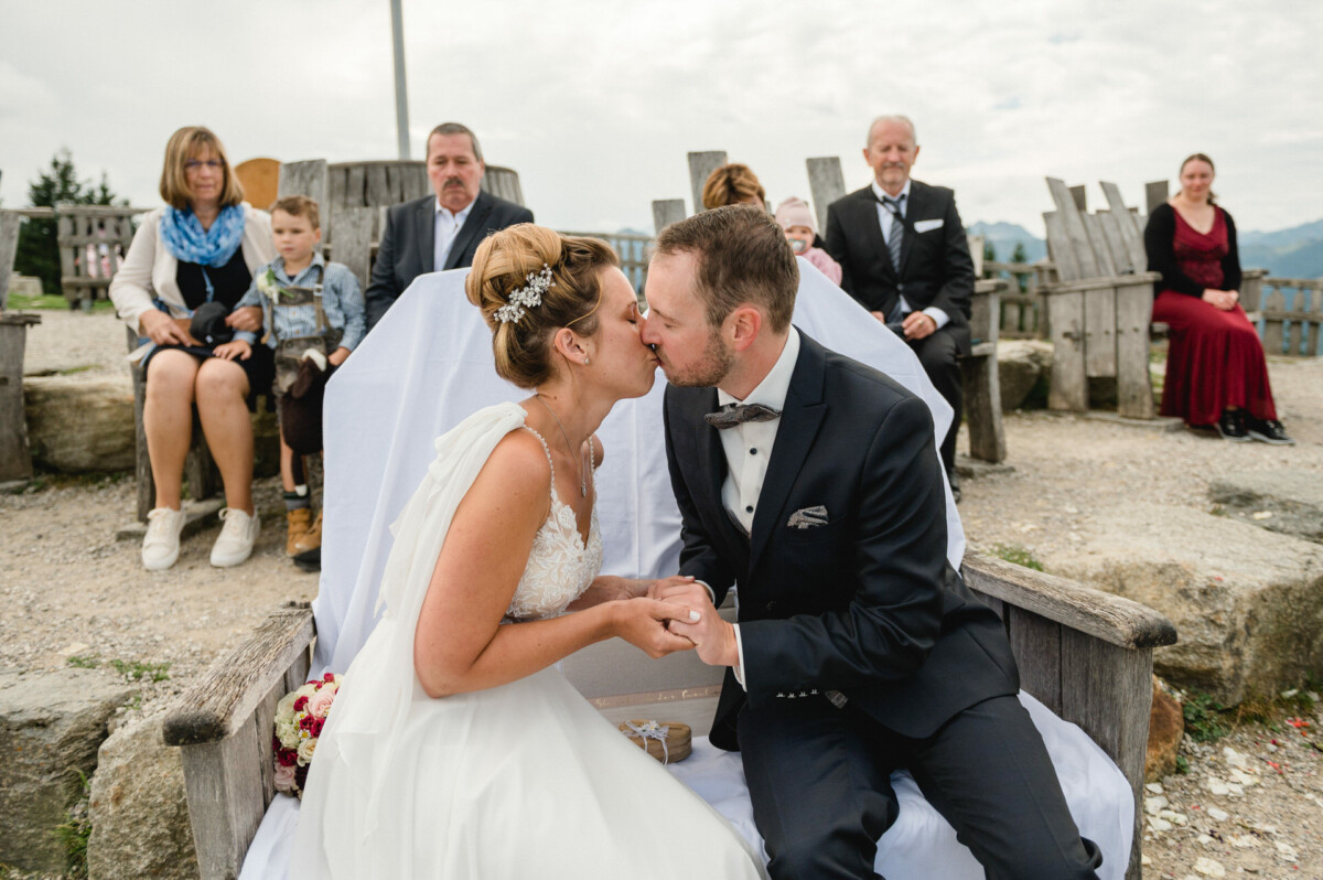 Braut und Bräutigam küssen sich, während sie im Freien auf Holzstühlen sitzen und von einer Gruppe Menschen umgeben sind.