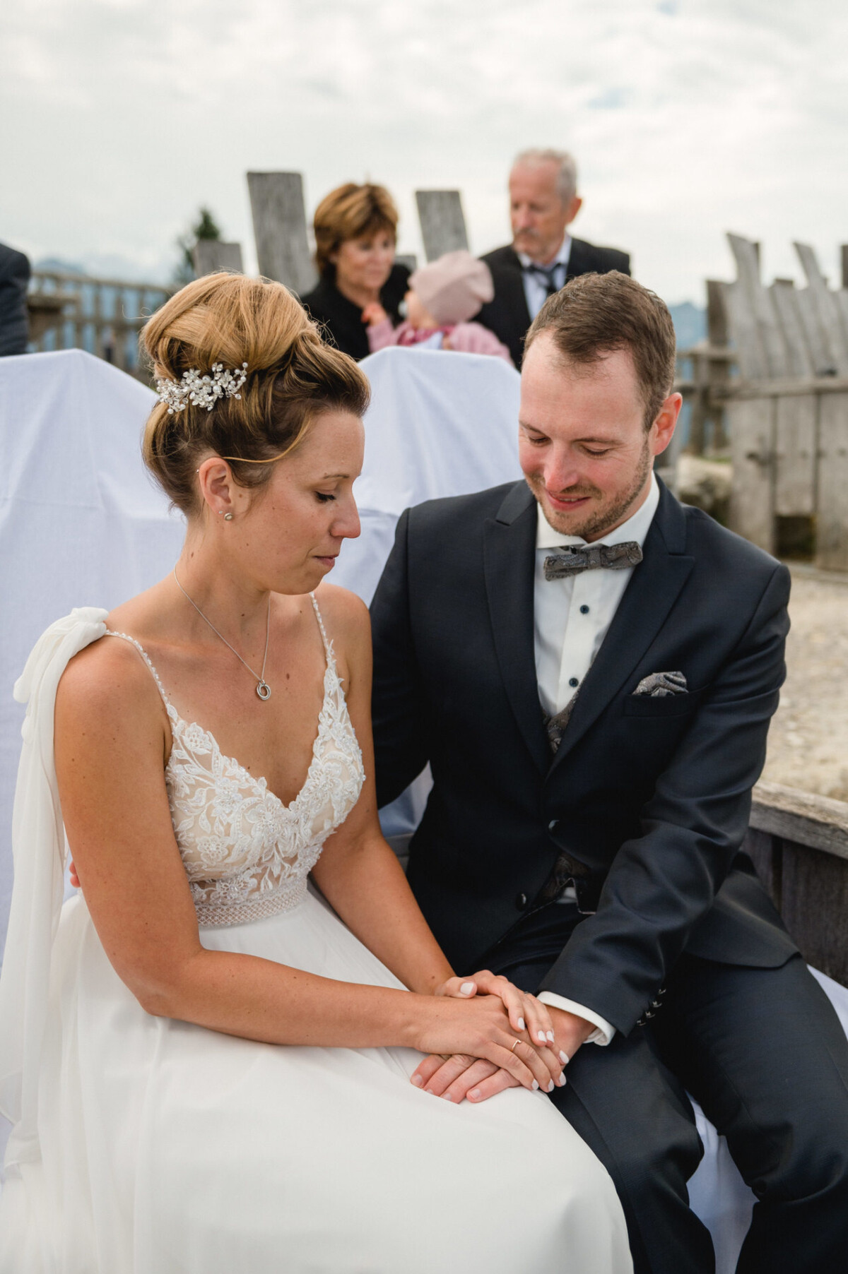 Braut und Bräutigam sitzen bei einer Hochzeitszeremonie im Freien eng beieinander und halten sich an den Händen. Sie wirken nachdenklich und in den Moment vertieft. Im Hintergrund sitzen zwei Personen.