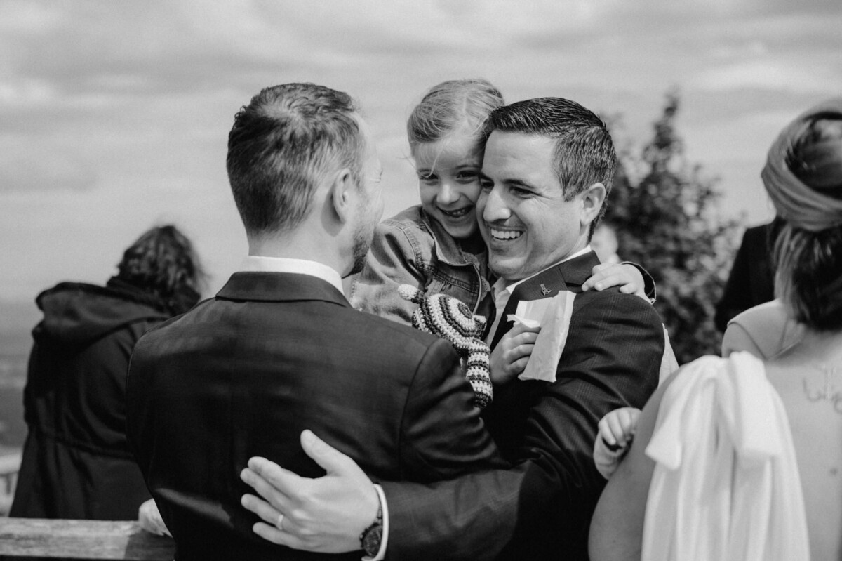 Zwei Männer lächeln und halten ein junges Mädchen im Freien. Eine Frau steht in der Nähe und im Hintergrund ist eine Person mit Kapuze zu sehen.