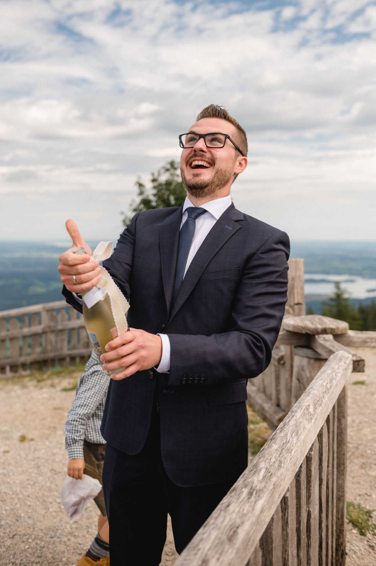 Ein Mann im Anzug öffnet freudig eine Flasche Champagner im Freien neben einem Holzzaun, im Hintergrund eine malerische Landschaft.