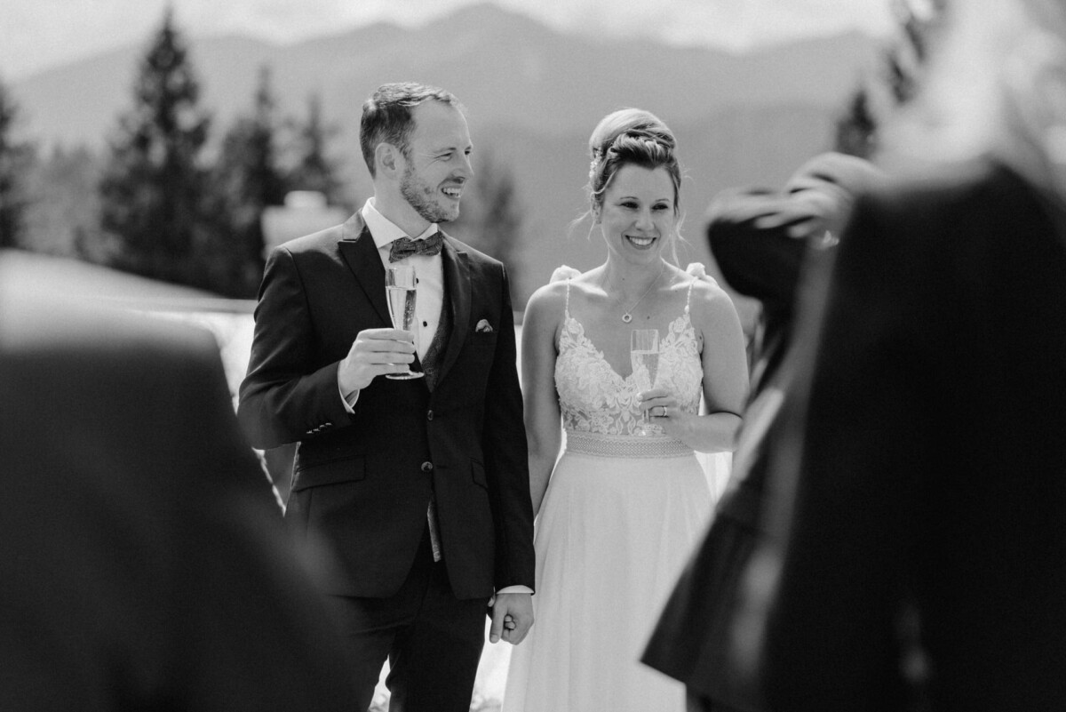 Braut und Bräutigam stehen im Freien und halten Champagnergläser in der Hand. Lächelnd. Im Hintergrund sind Bäume und Berge zu sehen. Schwarzweißbild.