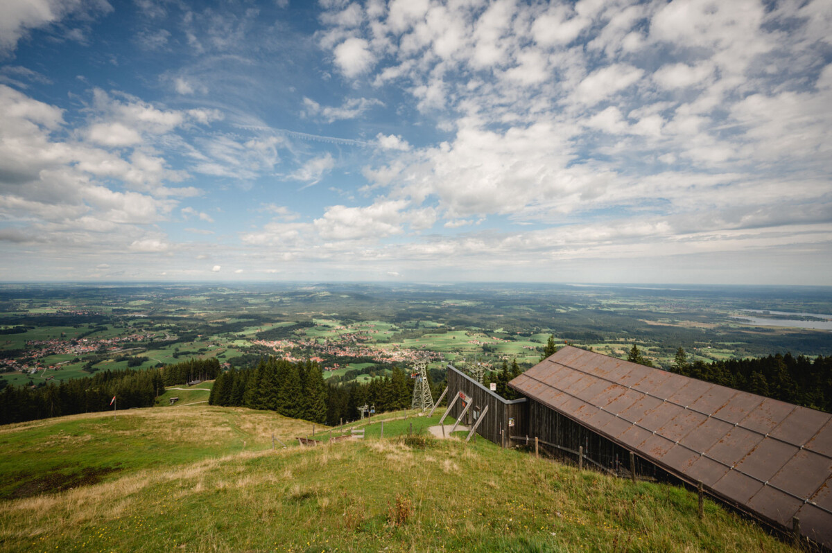 Eine malerische Aussicht von einem Hügel auf eine weite Landschaft mit Feldern, Wäldern und verstreuten Gebäuden unter einem teilweise bewölkten Himmel. Im Vordergrund ist eine Metalldachkonstruktion zu sehen.