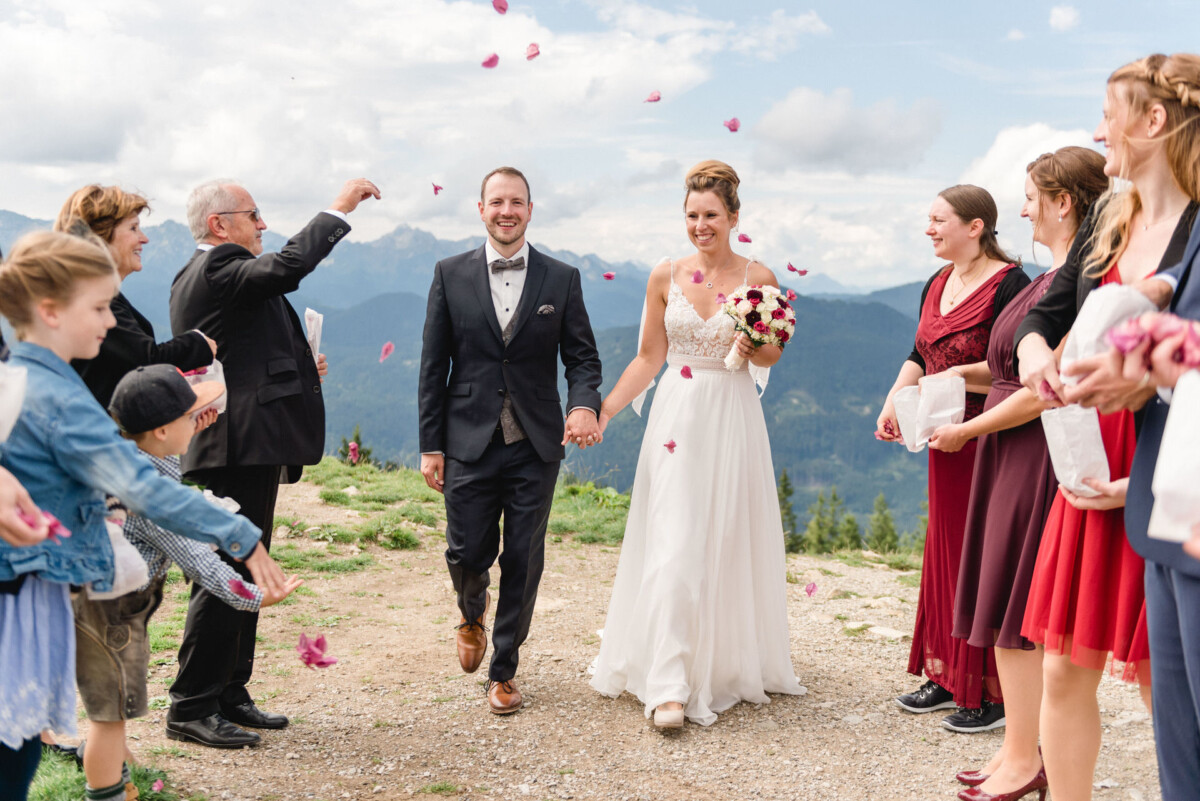 2022.09.03-Daniela-Benjamin-Hochzeit-Bad-Kohlgrub-am-Hoernle-Garmisch-Partenkirchen-00345 Braut und Bräutigam gehen Hand in Hand im Freien, während die Gäste Blütenblätter nach ihnen werfen. Im Hintergrund sind Berge zu sehen und die Gruppe lächelt.