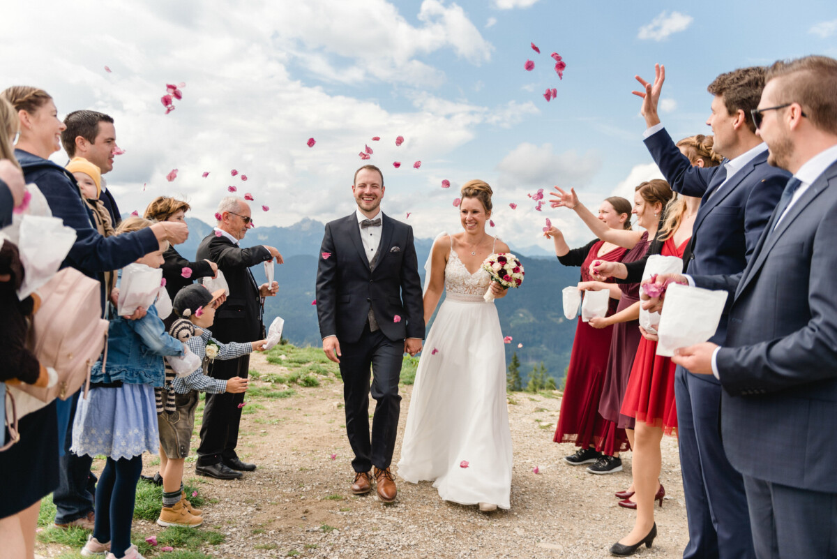 Braut und Bräutigam schreiten im Freien einen Gang entlang, während die Gäste aus feierlichen Gründen mit Blütenblättern werfen.