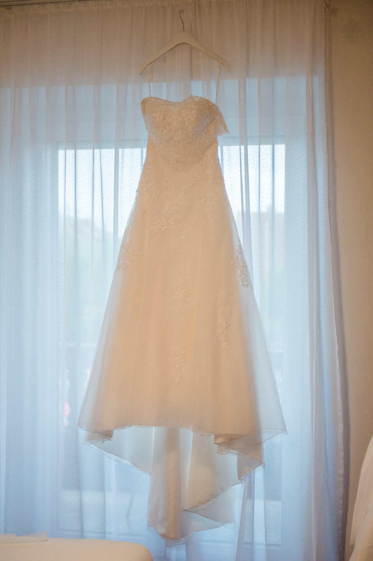 Ein weißes trägerloses Brautkleid hängt auf einem Kleiderbügel vor einem Fenster mit durchsichtigen Vorhängen.