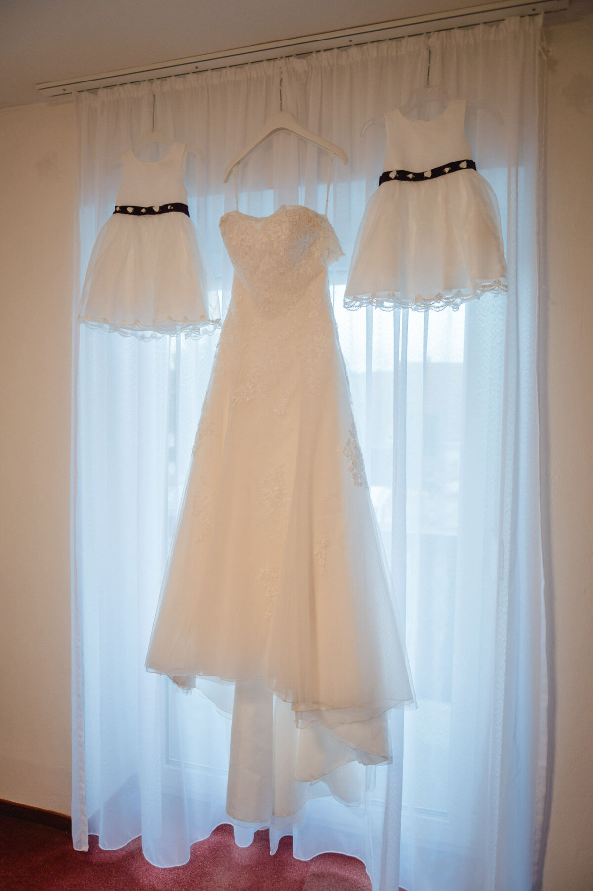 An einem Fenstervorhang hängen ein Brautkleid und zwei weiße Blumenmädchenkleider mit schwarzen Schärpen.