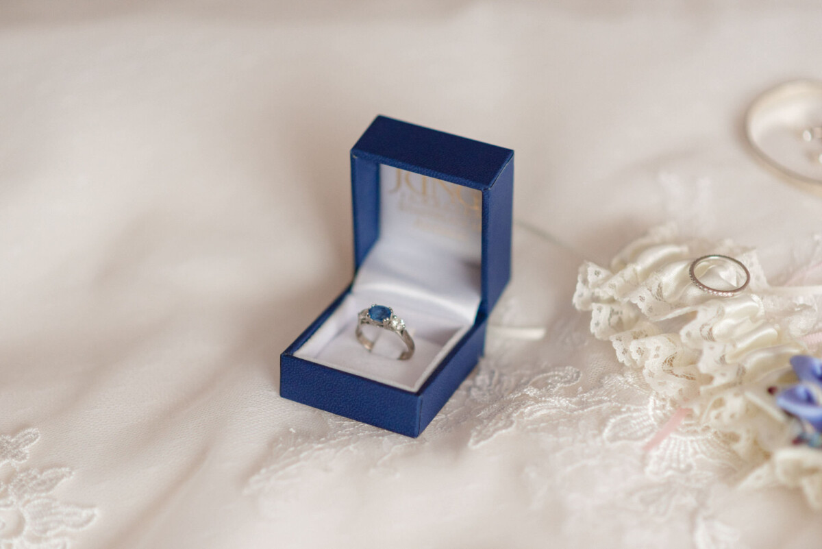 Eine blaue Ringschachtel enthält einen silbernen Ring mit einem blauen Edelstein und liegt geöffnet auf einem Spitzenstoff.