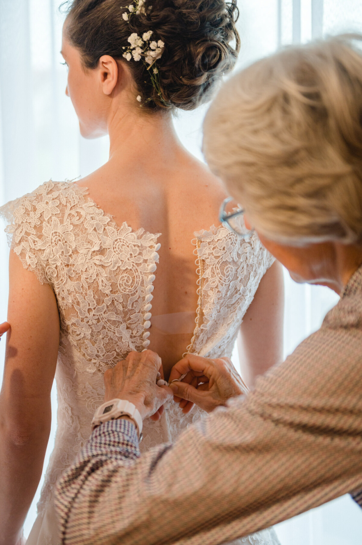 Eine ältere Frau hilft einer Braut in einem gut beleuchteten Raum, indem sie die Rückseite ihres Spitzenbrautkleides zuknöpft.