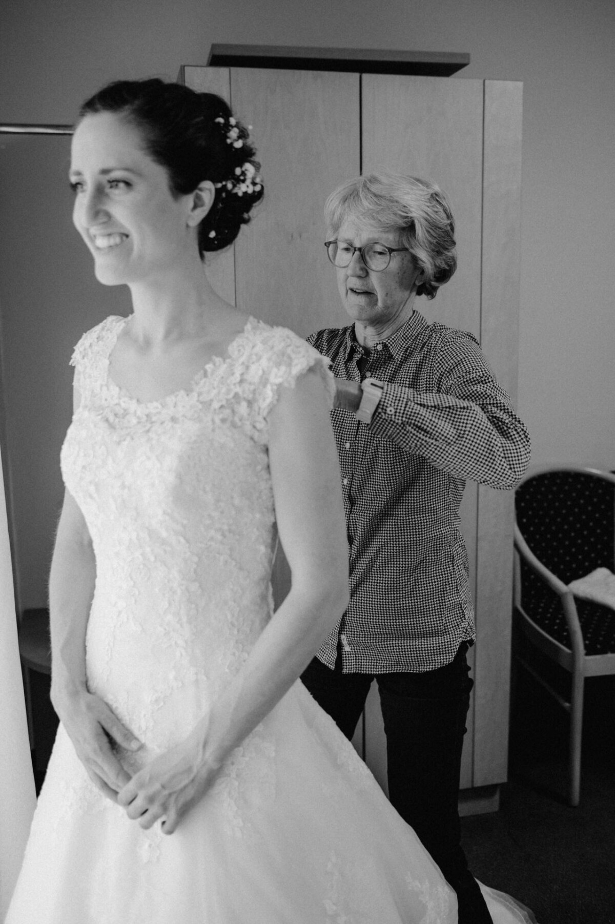 Einer Frau in einem Brautkleid wird in einem Raum von einer anderen Frau beim Zuschneiden des Rückens ihres Kleides geholfen.