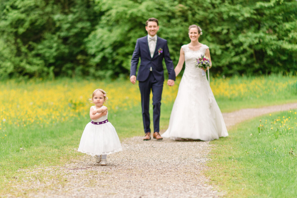 Ein junges Mädchen in einem weißen Kleid geht mit verschränkten Armen auf einem Kiesweg. Hinter ihr lächelt ein Paar in Hochzeitskleidung und hält sich an den Händen. Sie sind von Grünpflanzen und gelben Blumen umgeben.