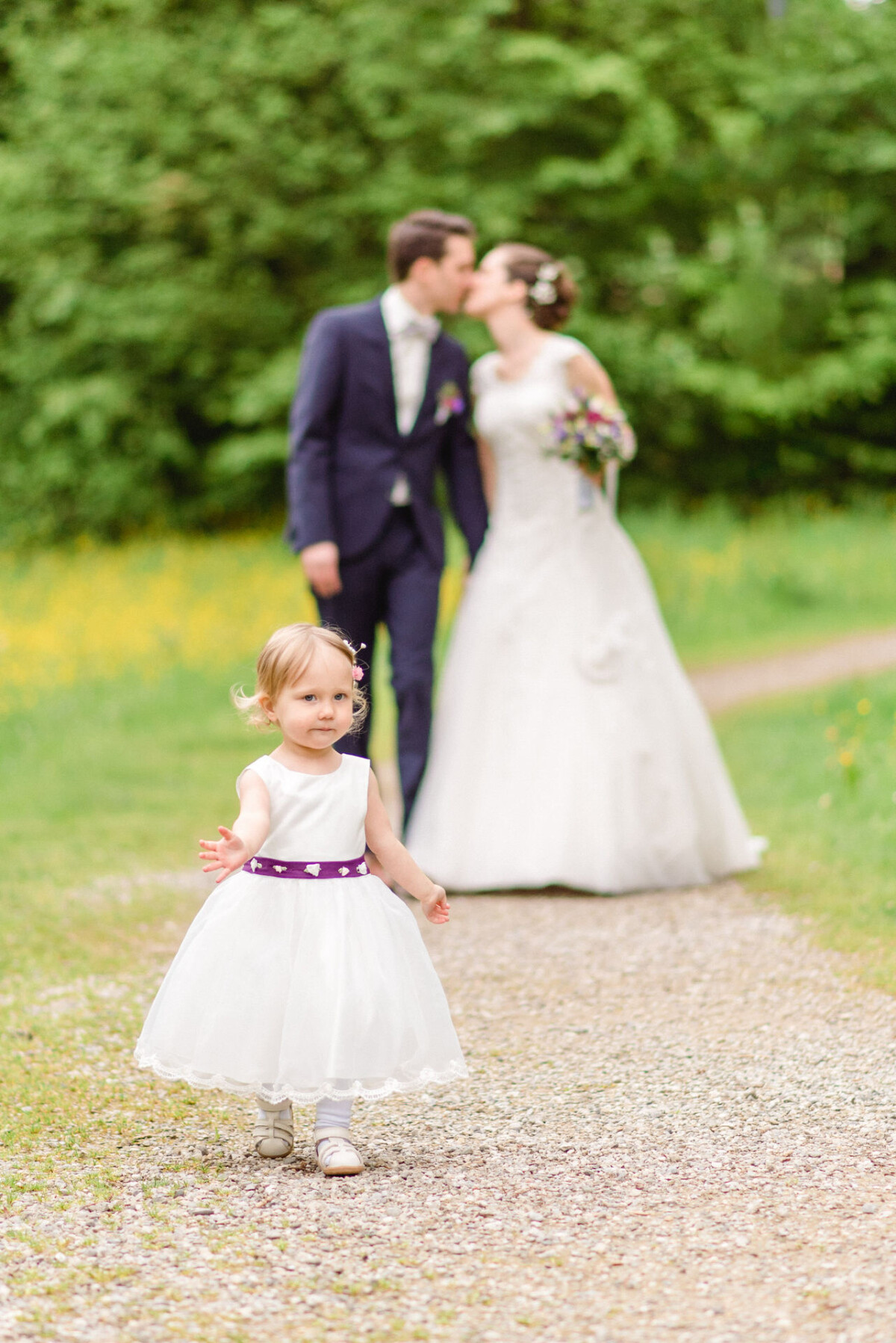 Ein Kleinkind im weißen Kleid läuft auf einem Kiesweg, während sich im unscharfen Hintergrund im Grünen Braut und Bräutigam küssen.