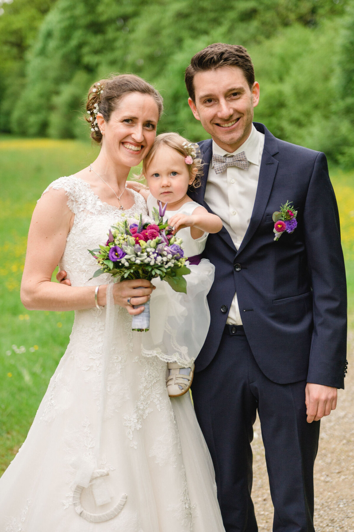 Ein Paar in Hochzeitskleidung steht mit einem kleinen Kind im Freien. Die Frau hält einen Blumenstrauß und das Kind wird zwischen ihnen gehalten. Im Hintergrund sind Grünpflanzen zu sehen.