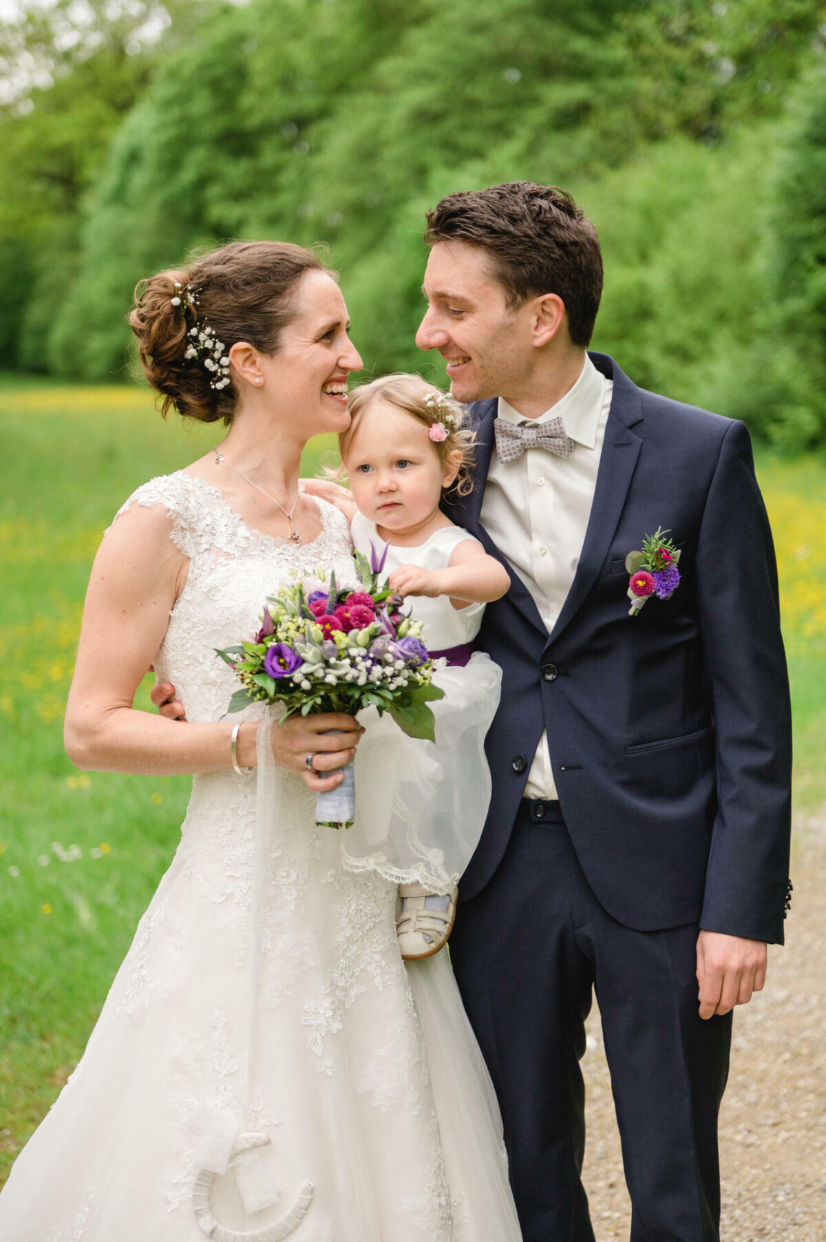 Braut und Bräutigam lächeln einander an, halten ein kleines Kind und posieren auf einem Grasweg. Die Braut trägt ein weißes Kleid mit Blumen und der Bräutigam einen Anzug mit Fliege.
