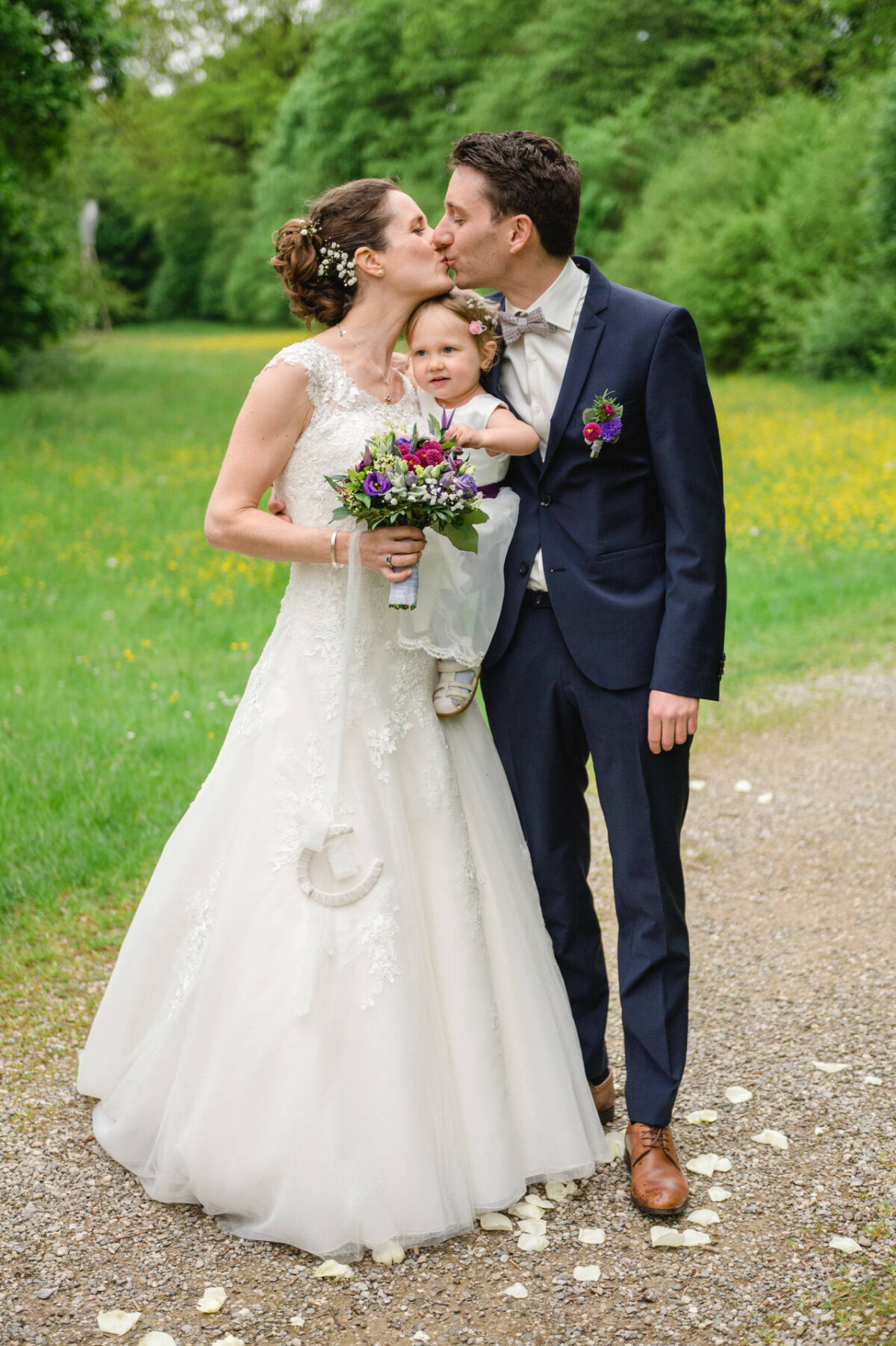 Braut und Bräutigam küssen sich und halten ein Baby auf einem von Grün gesäumten Weg. Die Braut hält einen Strauß lila Blumen.