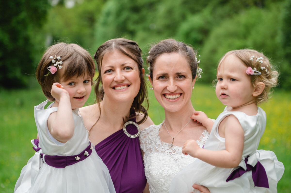 Zwei Frauen lächeln, während sie zwei junge Mädchen im Arm halten, auf einer Wiese im Freien. Die Frauen sind in Abendgarderobe gekleidet, und die Mädchen tragen weiße Kleider mit violetten Schärpen.