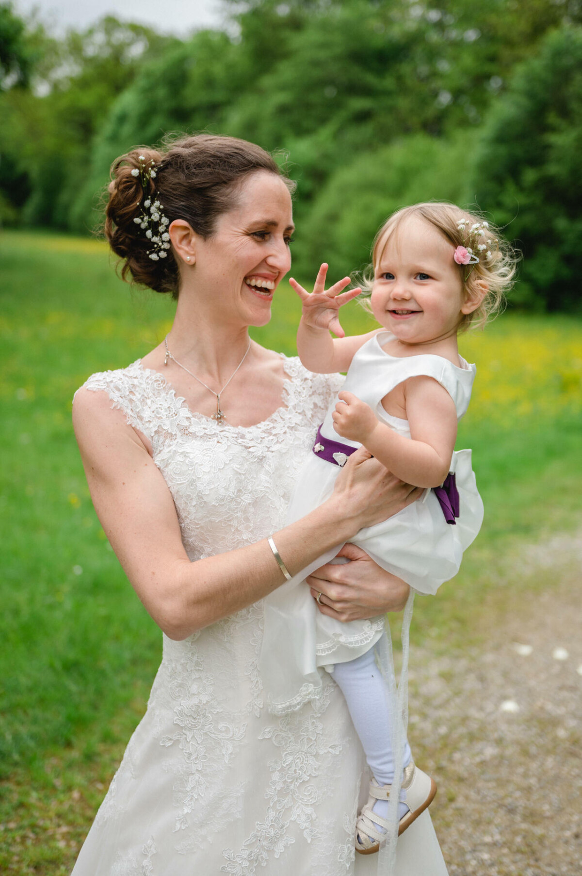 Eine Frau in einem weißen Kleid hält ein lächelndes Kleinkind mit Blumen im Haar und steht auf einem Grasweg mit Bäumen im Hintergrund.