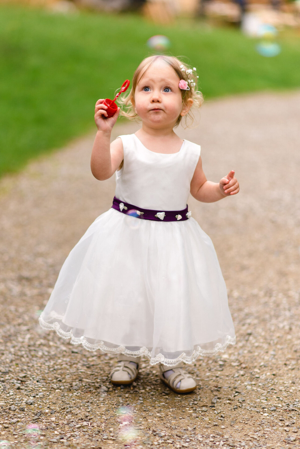 Ein junges Mädchen in einem weißen Kleid mit lila Gürtel steht auf einem Weg und hält einen roten Seifenblasenstab.