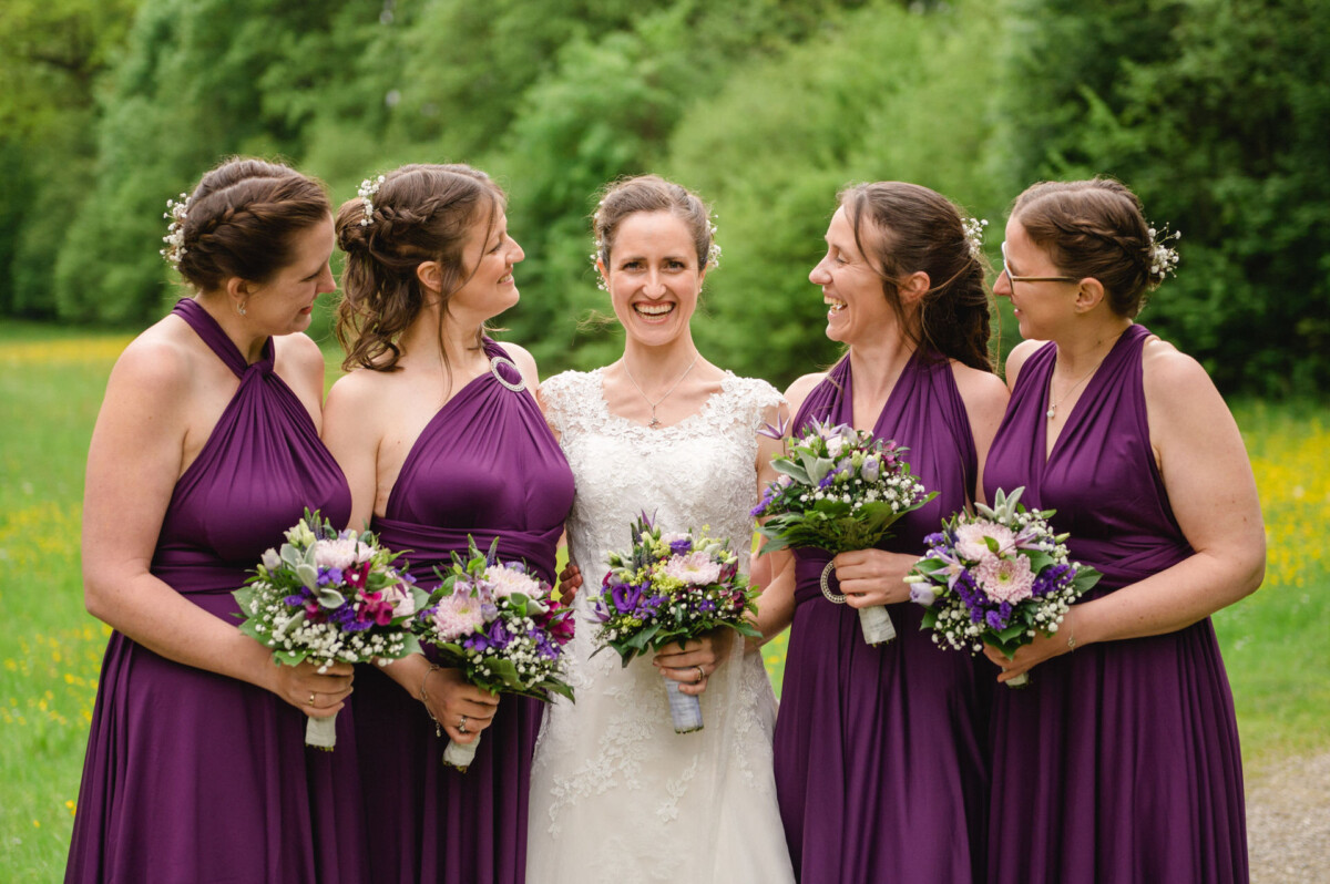 Eine Braut in einem weißen Kleid steht mit vier Brautjungfern in lila Kleidern, alle halten Blumensträuße, in einer üppigen Umgebung im Freien.