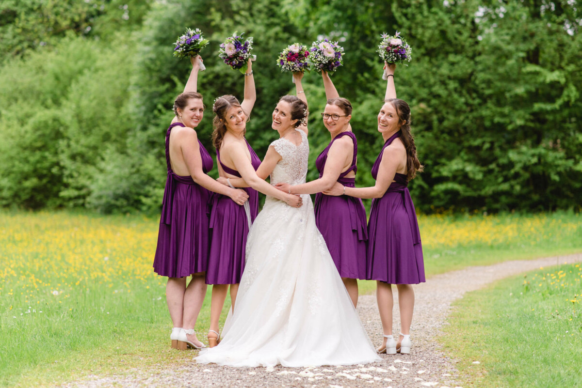 Eine Braut im weißen Kleid posiert auf einem Weg mit fünf Brautjungfern in lila Kleidern, die Blumensträuße halten, umgeben von Grün.