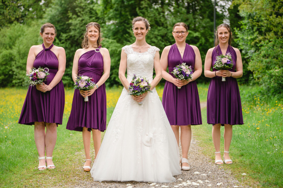 Eine Braut in einem weißen Kleid steht mit vier Brautjungfern in lila Kleidern, von denen jede einen Blumenstrauß hält, auf einem Weg in einer Rasenfläche mit Bäumen im Hintergrund.