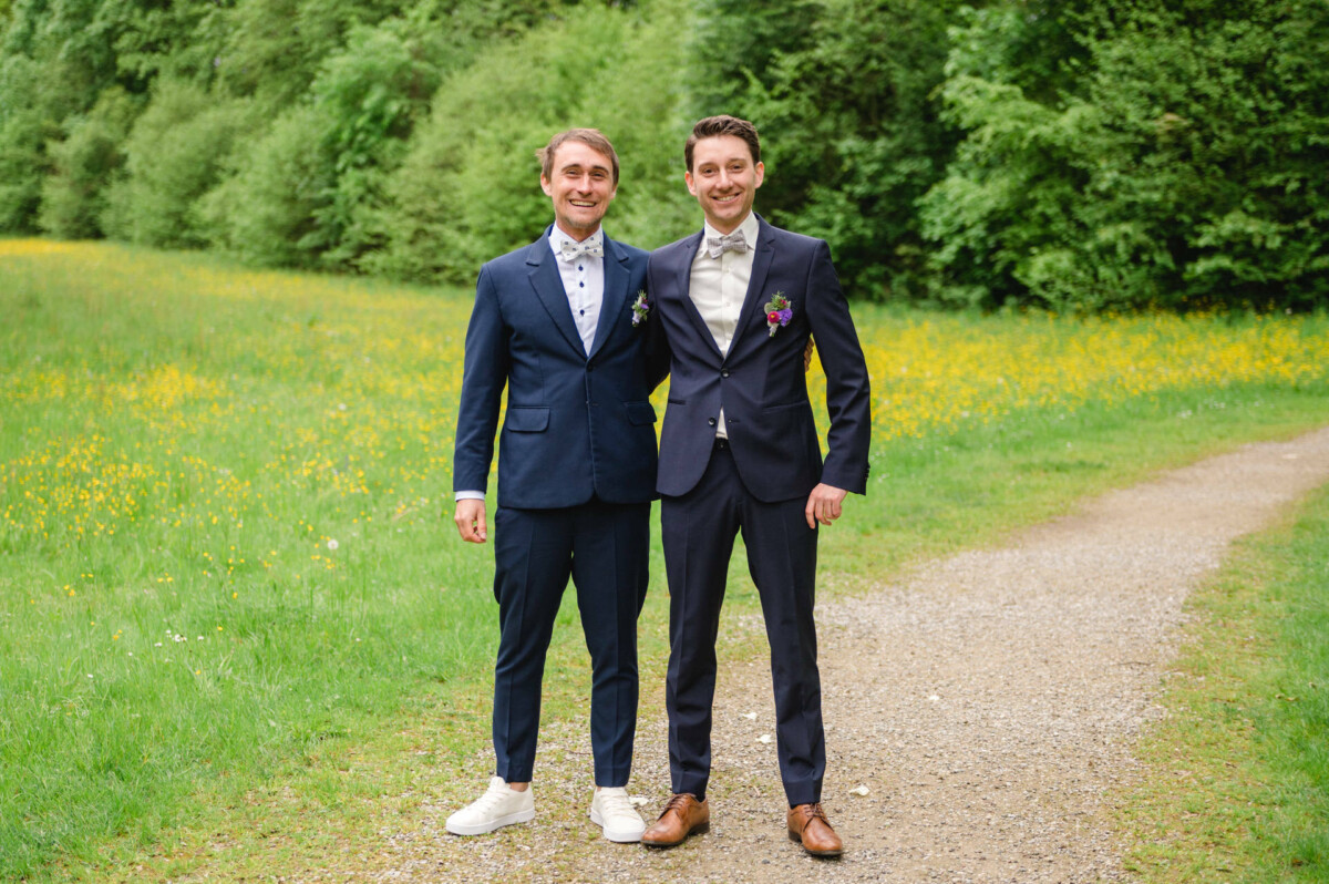 Zwei Männer in Anzügen stehen lächelnd auf einem Kiesweg in einer grasbewachsenen Waldgegend mit gelben Blumen.