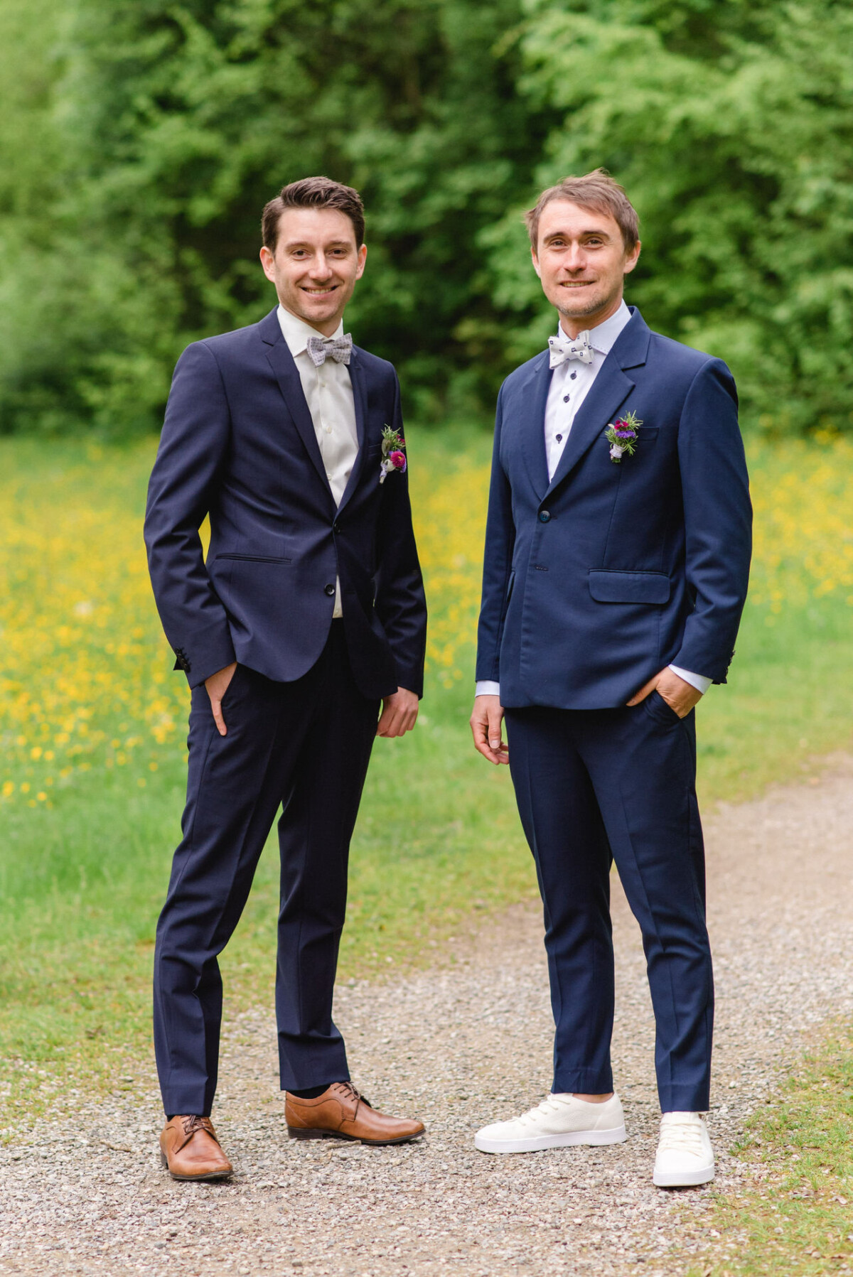 Zwei Männer in Anzügen stehen auf einem Kiesweg in einem grünen Park mit gelben Blumen im Hintergrund.
