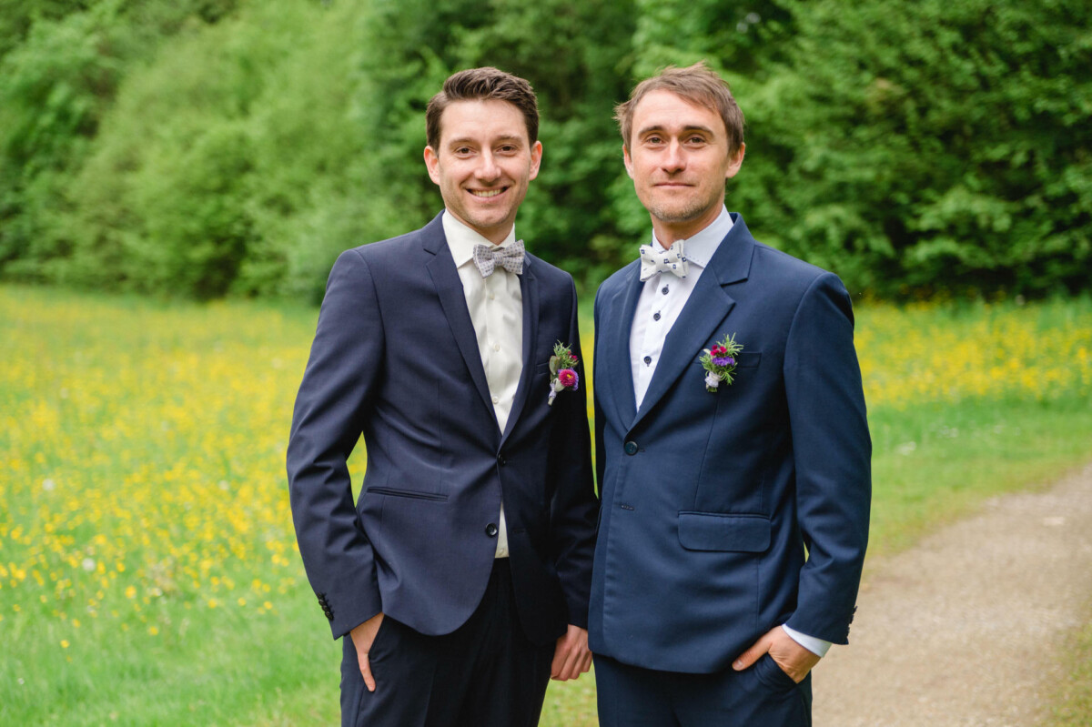 Zwei Männer in Anzügen stehen auf einem Weg in einem Feld mit grünen Bäumen und gelben Blumen im Hintergrund. Beide tragen Fliegen und Blumenboutonnieren.
