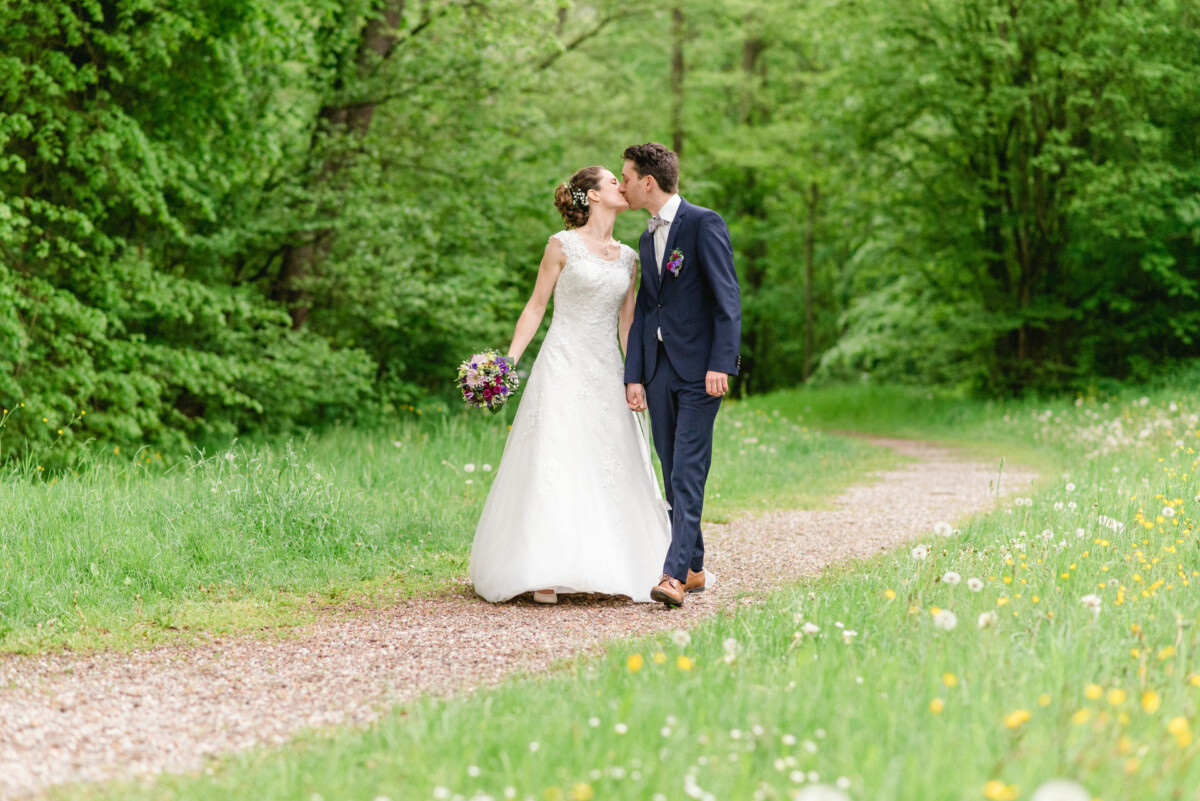 Braut und Bräutigam gehen einen Waldweg entlang, umgeben von Grün und Wildblumen. Die Braut hält einen Blumenstrauß und es sieht so aus, als würden sie sich küssen.