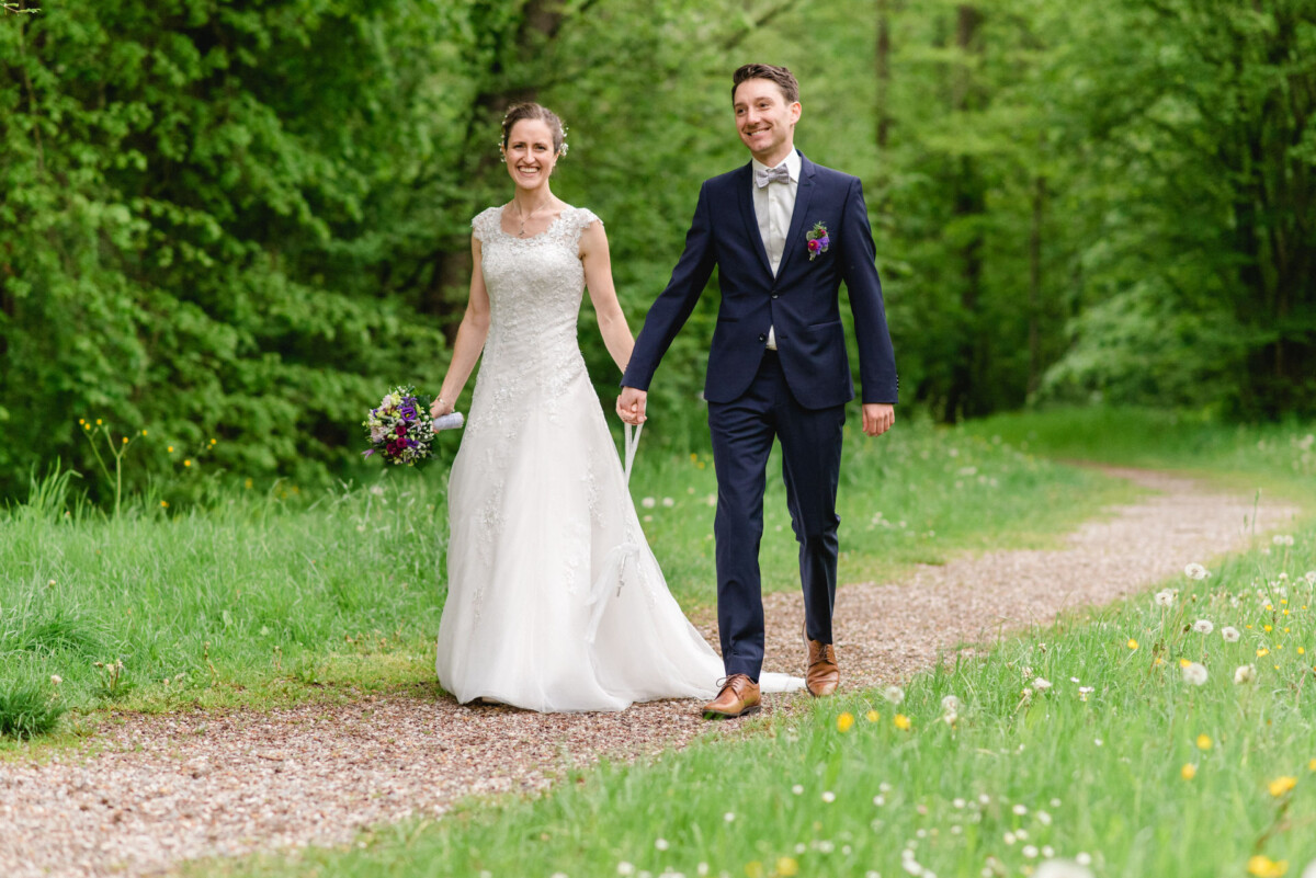 Braut und Bräutigam gehen Hand in Hand auf einem Waldweg, umgeben von üppigem Grün und Wildblumen.