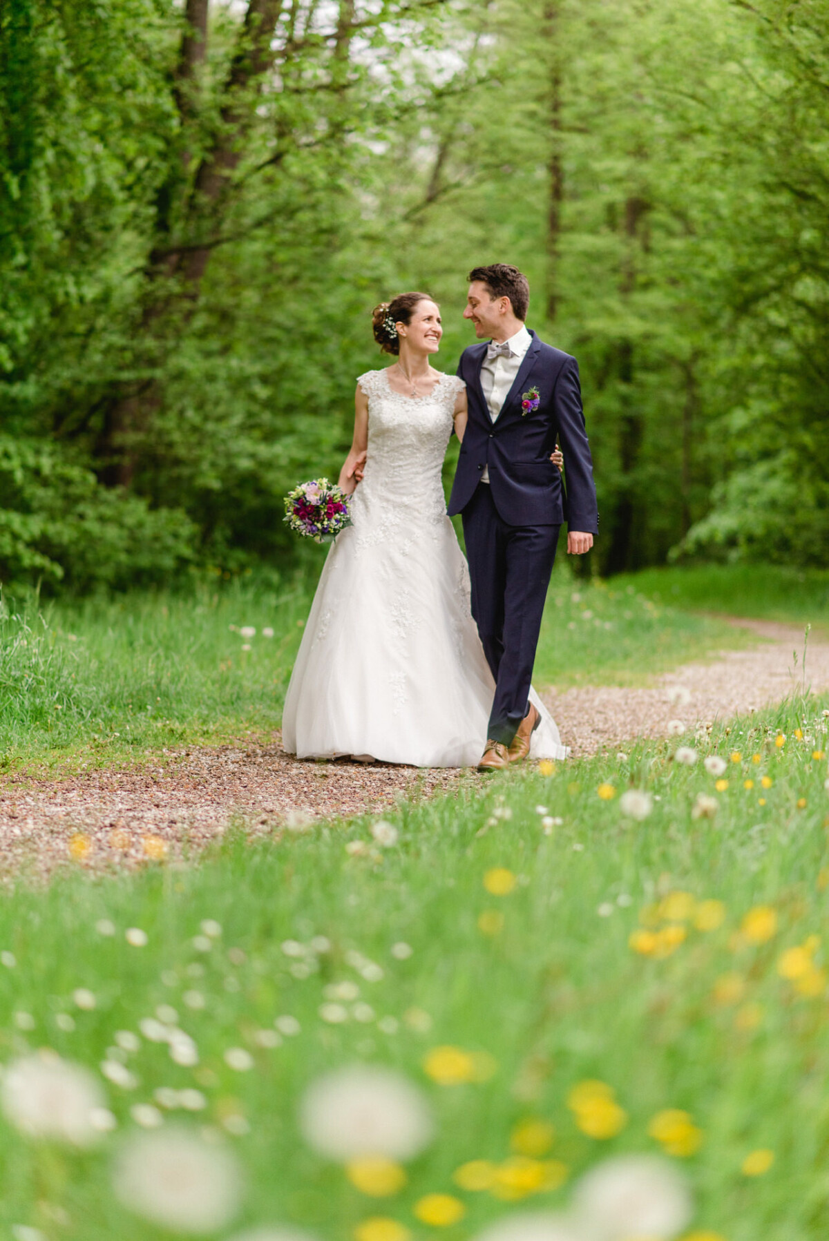 Eine Braut und ein Bräutigam im Hochzeitskleid gehen Arm in Arm auf einem Kiesweg, umgeben von grünen Bäumen und Wildblumen.