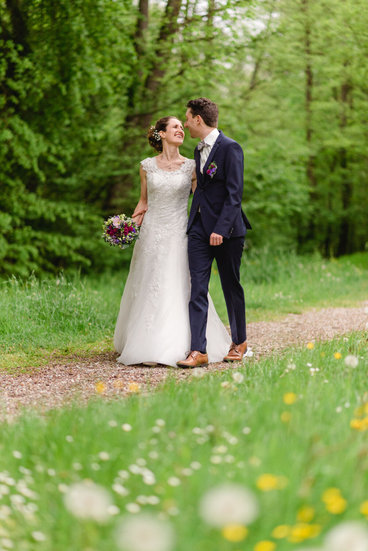 Ein Paar im Hochzeitskleid geht einen von grünen Bäumen umgebenen Grasweg entlang. Die Frau hält einen Blumenstrauß in der Hand.