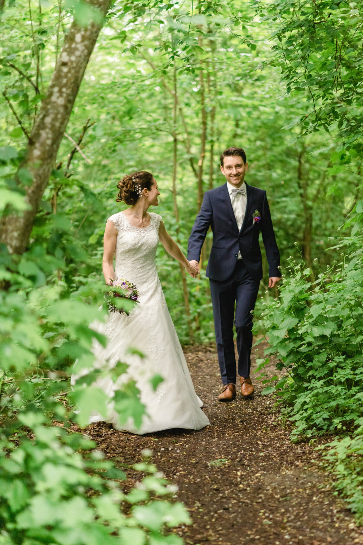 Braut und Bräutigam gehen Händchen haltend auf einem Waldweg, umgeben von grünem Laub. Die Braut trägt ein weißes Kleid und der Bräutigam einen marineblauen Anzug.