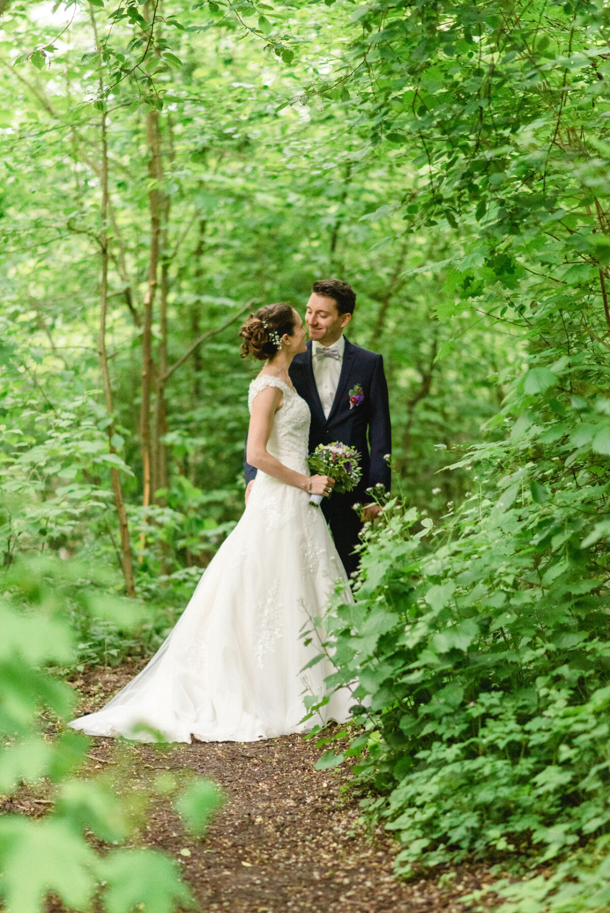 Braut und Bräutigam stehen zusammen in einem Waldgebiet, umgeben von üppigem Grün. Die Braut trägt ein weißes Kleid und hält Blumen, während der Bräutigam einen dunklen Anzug mit Fliege trägt.