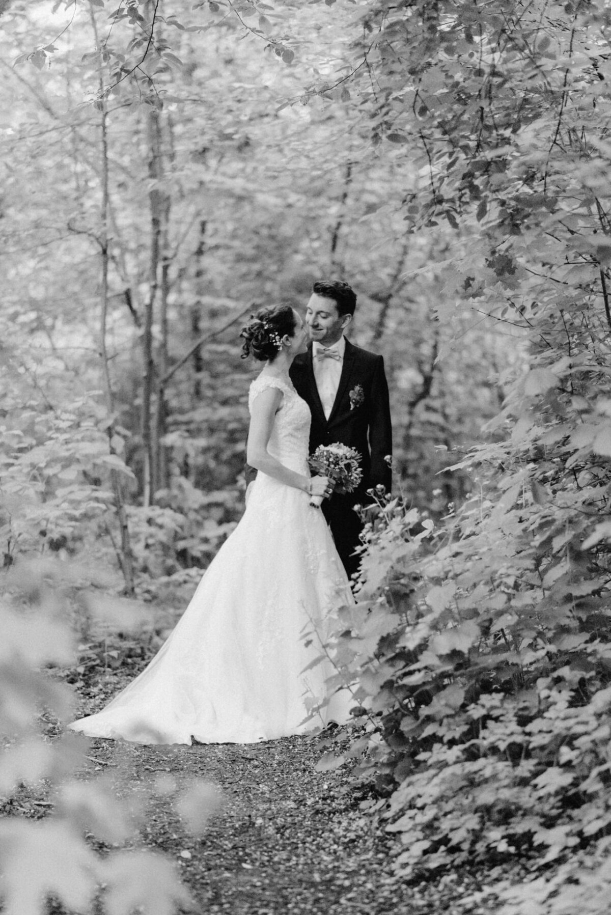 Braut und Bräutigam stehen auf einem Waldweg, umgeben von Bäumen. Die Braut trägt ein weißes Kleid und hält einen Blumenstrauß, und der Bräutigam trägt einen dunklen Anzug.