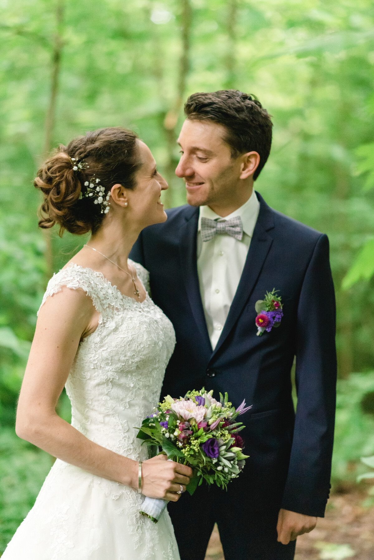 Braut und Bräutigam stehen in einem Wald und lächeln einander an. Die Braut hält einen Blumenstrauß in der Hand und trägt ein weißes Spitzenkleid, und der Bräutigam trägt einen marineblauen Anzug mit einer Blumenboutonniere.