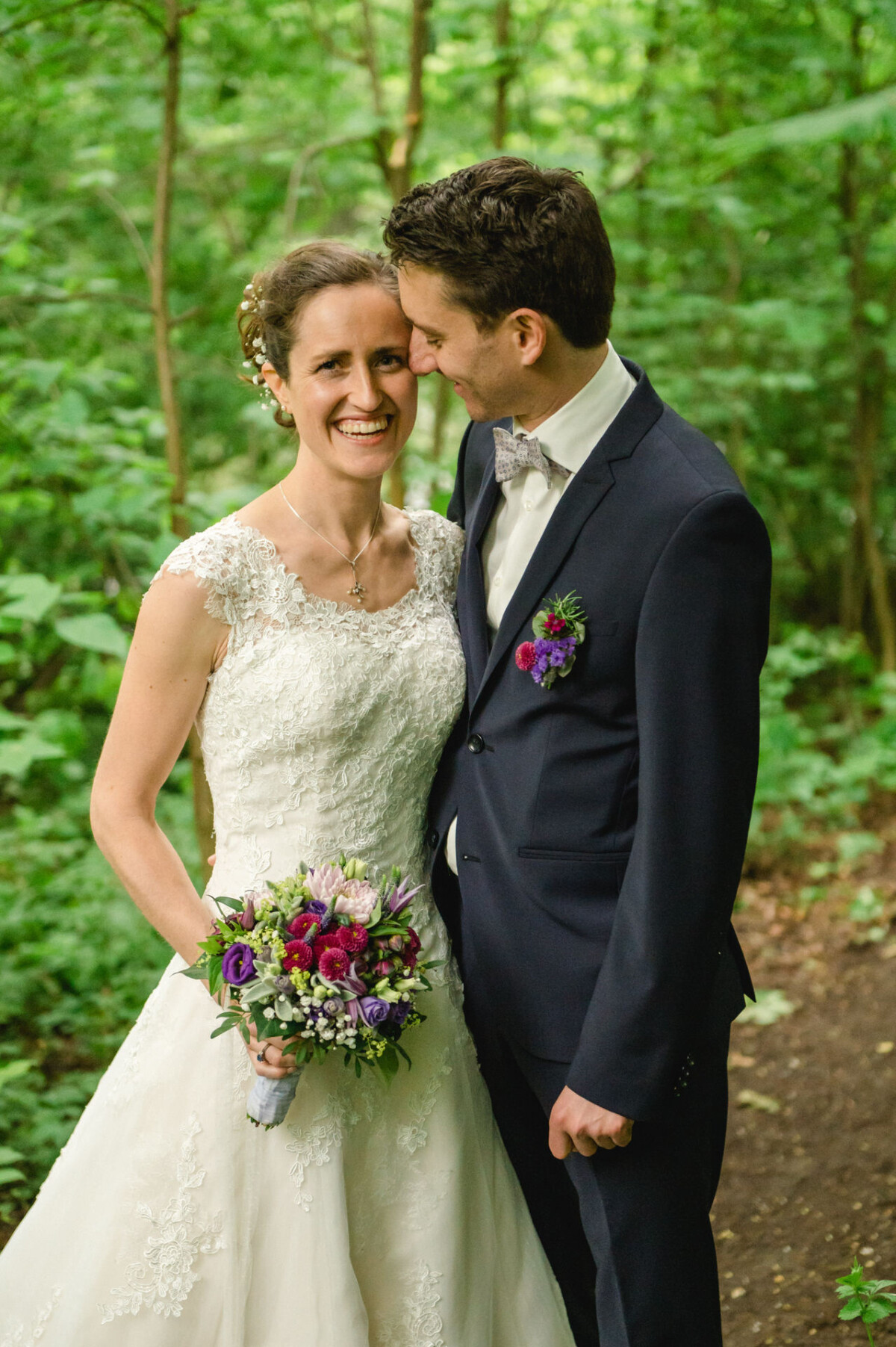 Eine Braut in einem Spitzenbrautkleid hält einen Blumenstrauß und steht neben einem Bräutigam im Anzug mit einer Blumenansteckblume, umgeben von Grün.