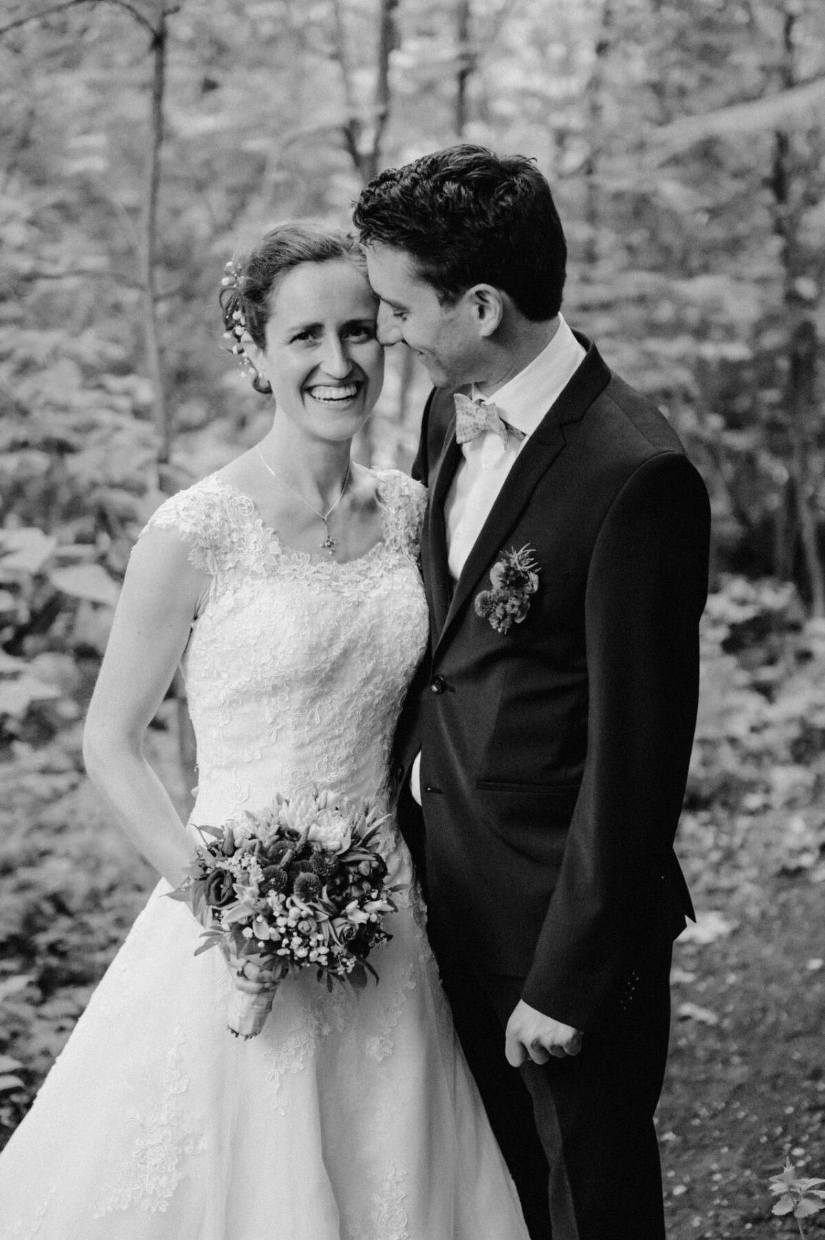 Braut und Bräutigam lächeln sich in einem Waldstück an, die Braut hält einen Blumenstrauß.