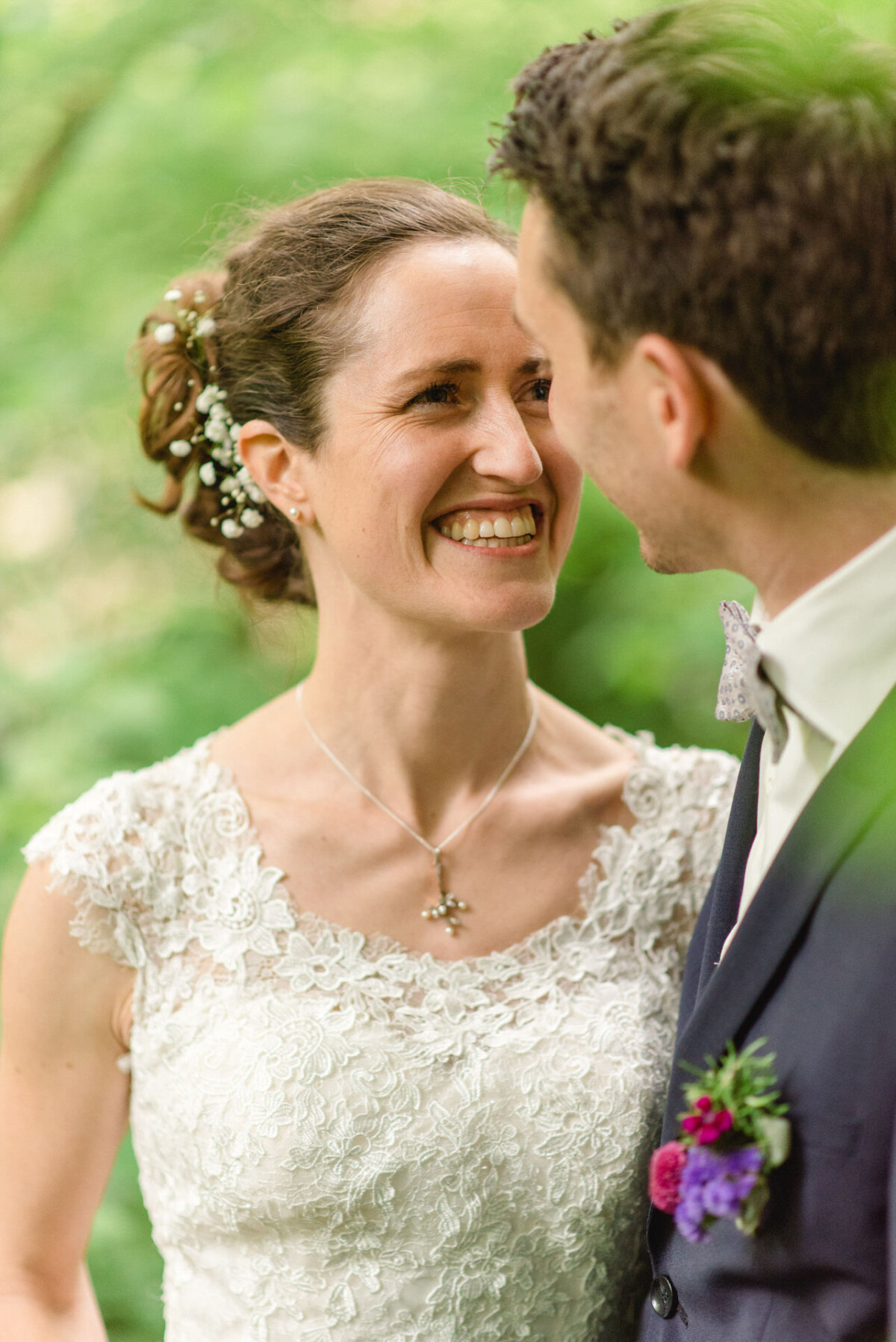 Braut und Bräutigam lächeln sich in einer Gartenkulisse an. Die Braut trägt ein weißes Spitzenkleid mit floralem Haarschmuck, der Bräutigam trägt einen Anzug mit einer Blumenboutonniere.