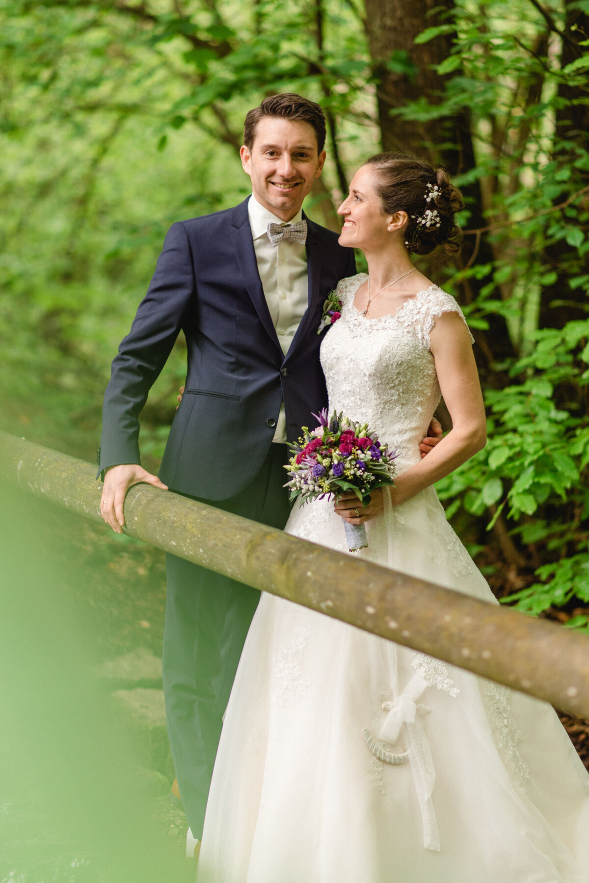 Braut und Bräutigam stehen auf einer Holzbrücke in einem Wald. Die Braut hält einen Strauß lila Blumen und beide lächeln.