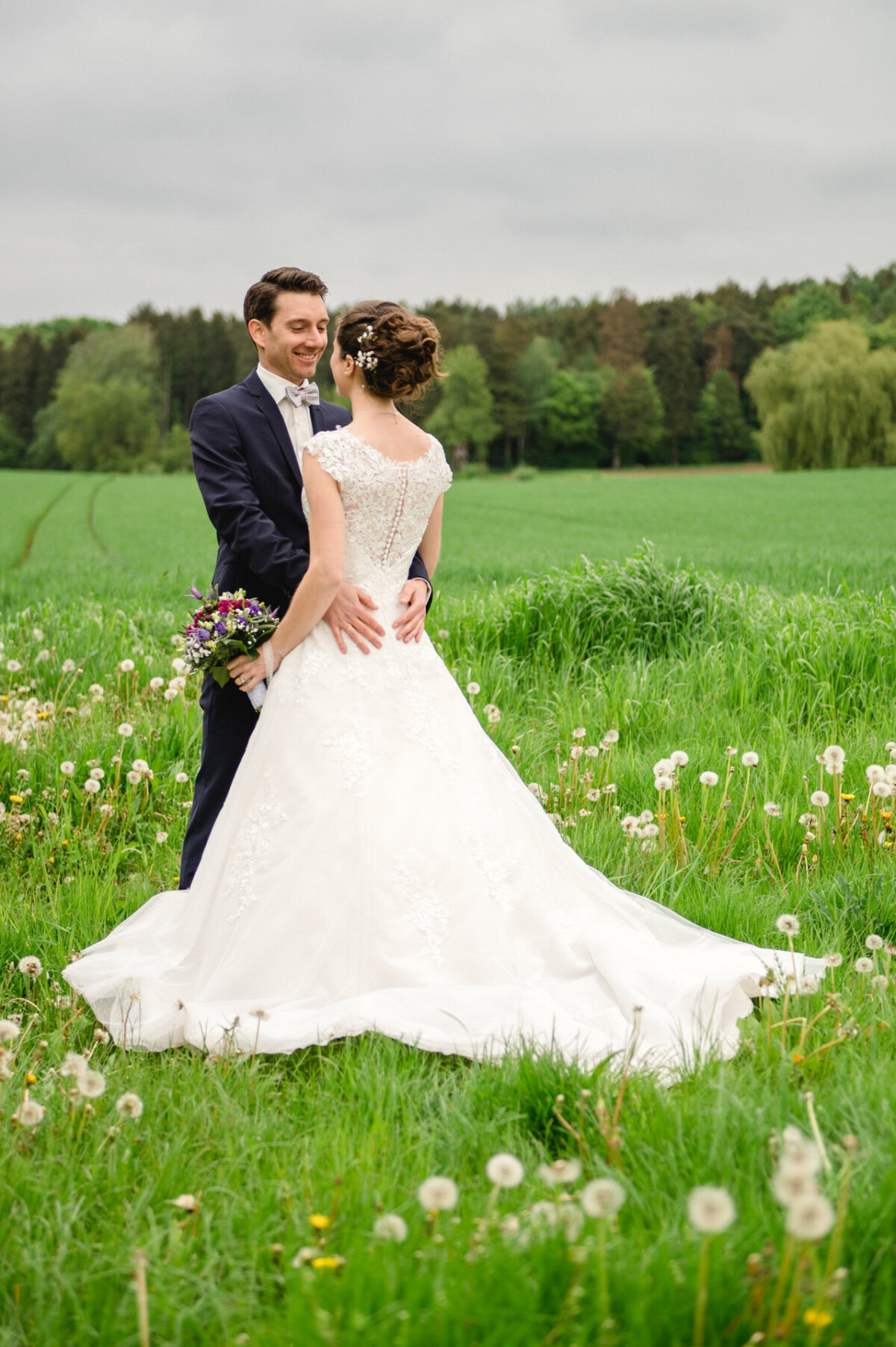 Braut und Bräutigam stehen auf einer grünen Wiese mit Bäumen im Hintergrund. Die Braut hält einen Blumenstrauß und trägt ein weißes Kleid, während der Bräutigam einen dunklen Anzug trägt.