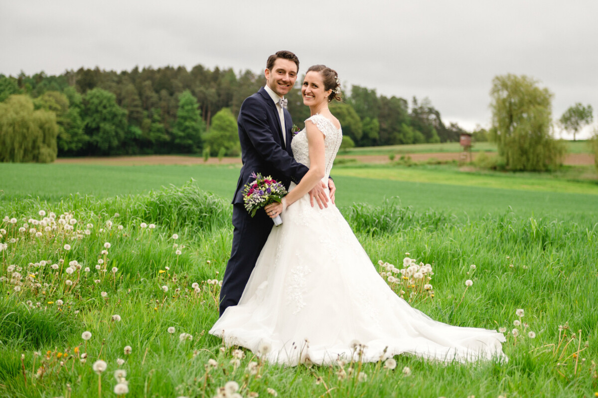 Ein Paar in Brautkleid und Anzug steht auf einer grünen Wiese mit Bäumen im Hintergrund.