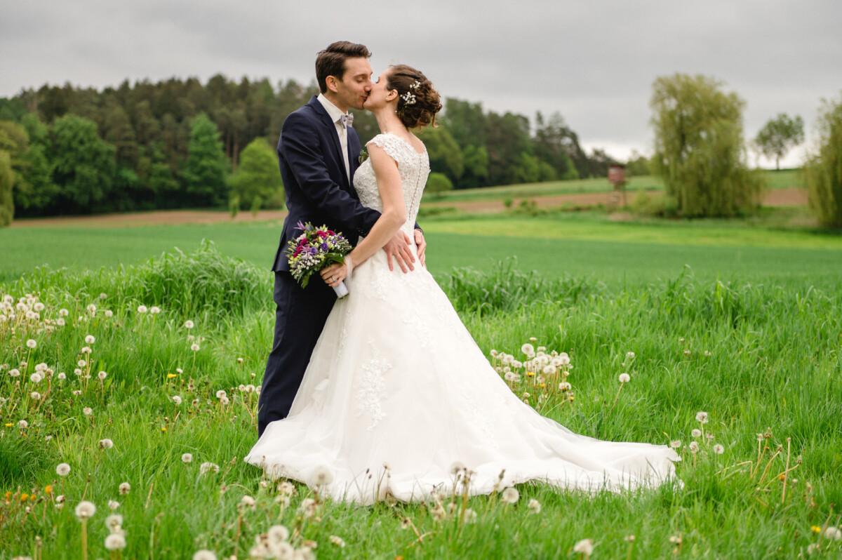 Braut und Bräutigam küssen sich auf einer grünen Wiese, umgeben von Bäumen und Löwenzahn.