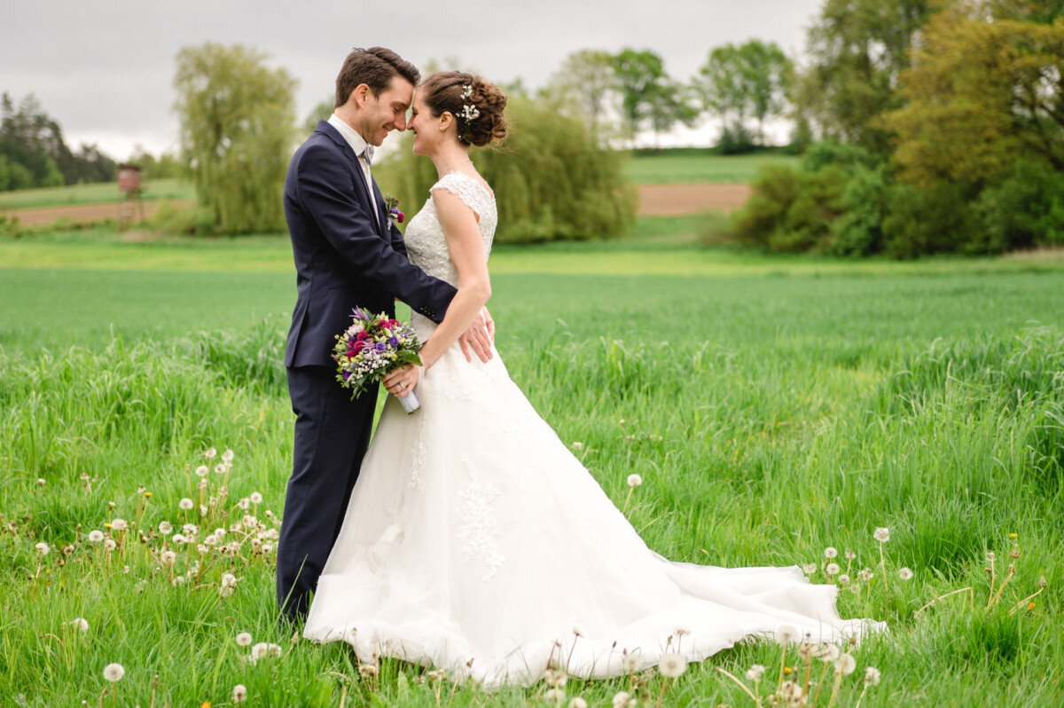 Braut und Bräutigam stehen eng beieinander auf einer grünen Wiese und lächeln einander an. Die Braut hält einen Blumenstrauß und beide tragen Hochzeitskleidung. Im Hintergrund sind Bäume zu sehen.