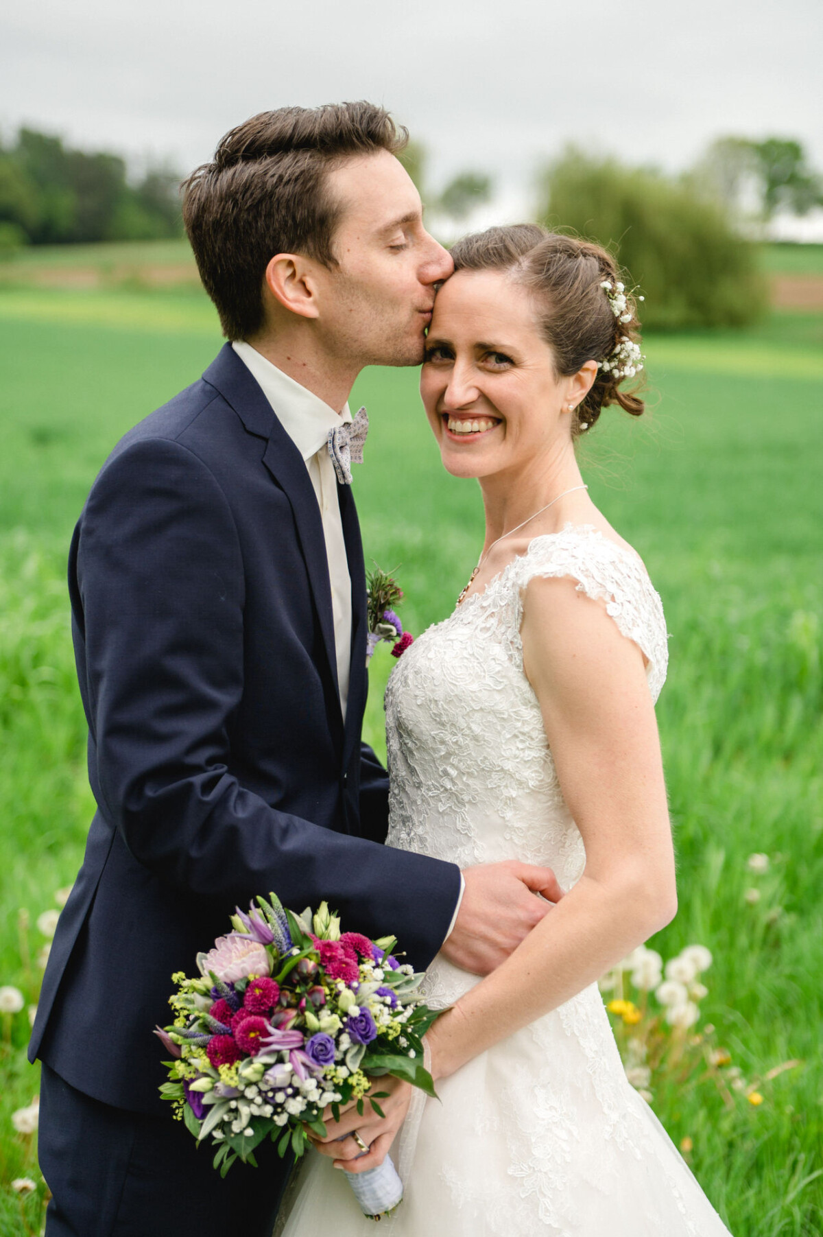 Ein Bräutigam im Anzug küsst die Stirn seiner lächelnden Braut, während sie auf einer grünen Wiese stehen; sie hält einen bunten Blumenstrauß.
