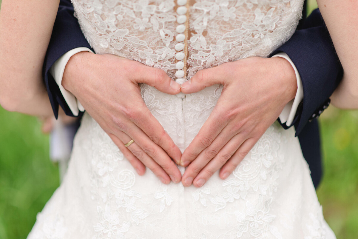 Nahaufnahme eines sich umarmenden Paares, dessen Hände über dem Rücken eines Spitzenbrautkleides ein Herz formen.